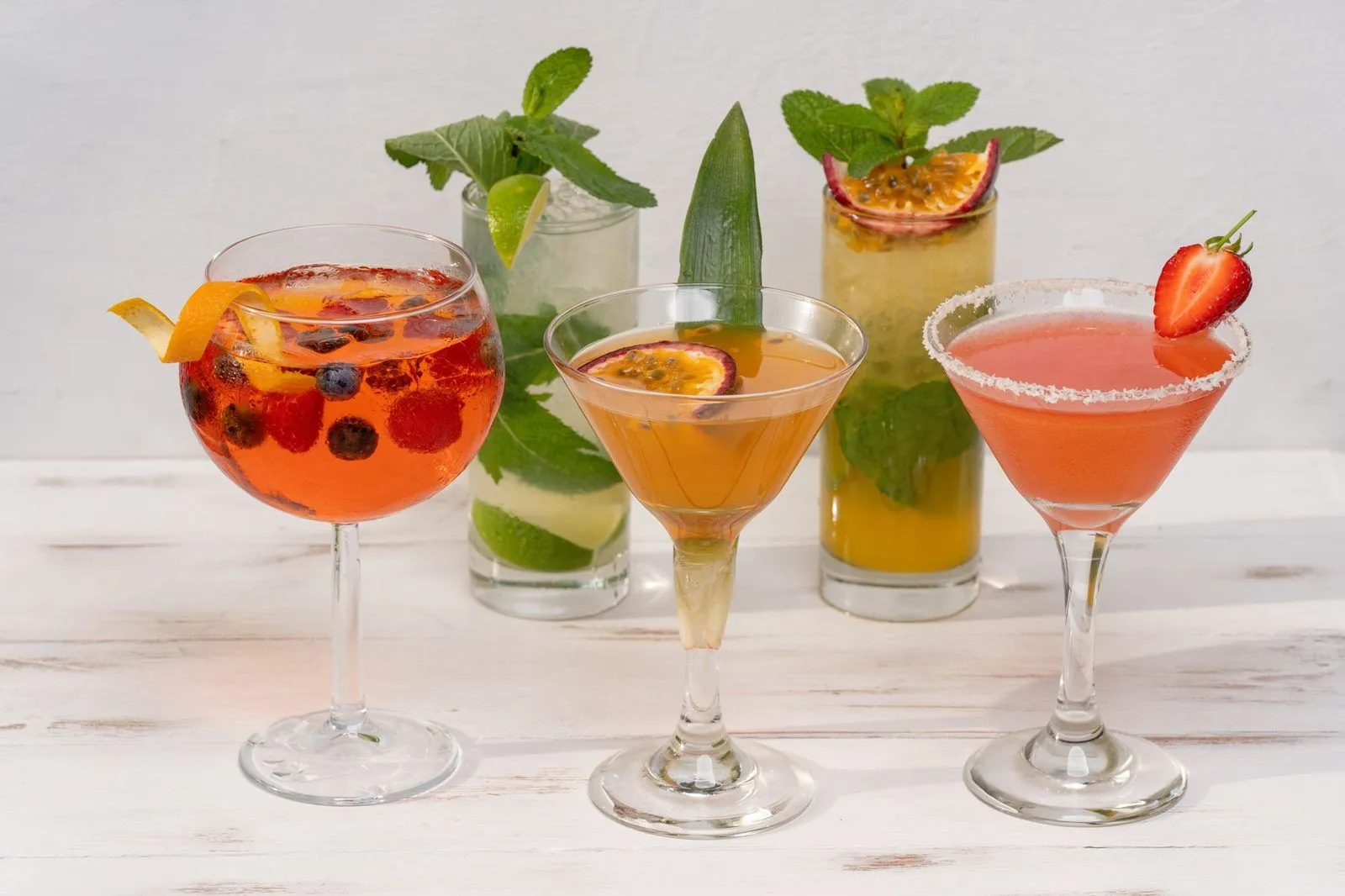 Cinco tragos coloridos sobre una mesa blanca: adelante, una copa con un trago anaranjado, arándanos, frambuesas y cáscara de naranja