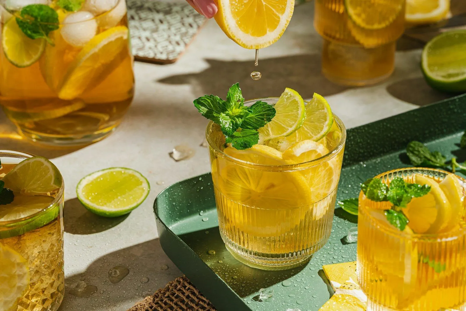 Mano exprimiendo medio limón sobre un vaso con hielo, rodajas de limón y lima y una ramita de menta