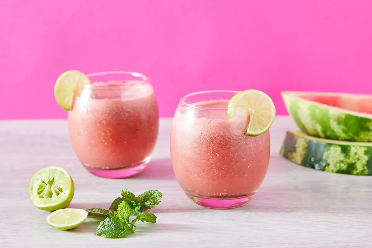 Dos vasos con granizado de sandía decorados con rodajas de lima, junto a menta fresca y rodajas de sandía