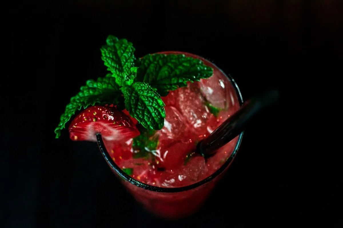Vaso de trago rojo de frutilla con hielo, hojas de menta y una frutilla en el borde, visto desde arriba sobre fondo oscuro