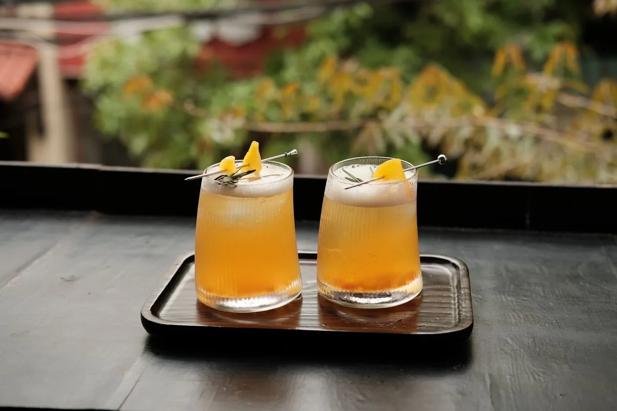 Dos vasos con un trago dorado y espumoso, decorados con trozos de fruta amarilla y romero en un palillo, sobre una bandeja de madera