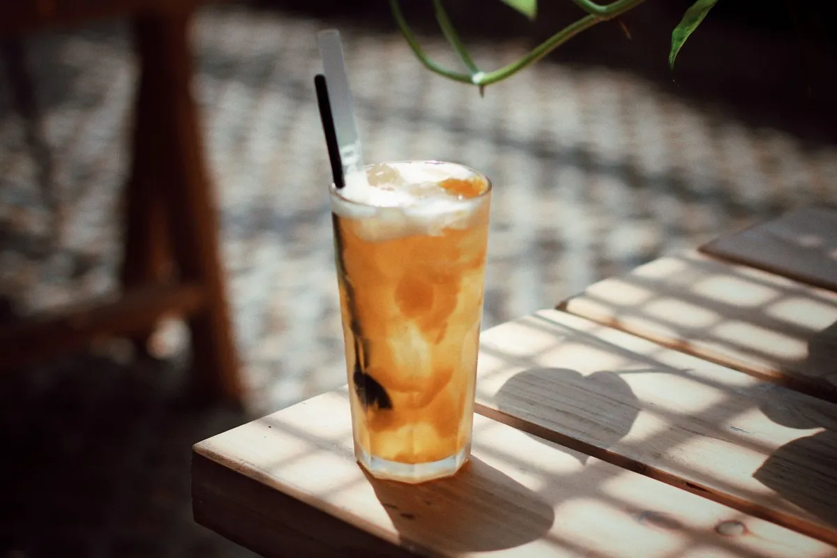 Vaso alto de té helado de durazno con hielo y trozos de fruta sobre una mesa de madera al sol